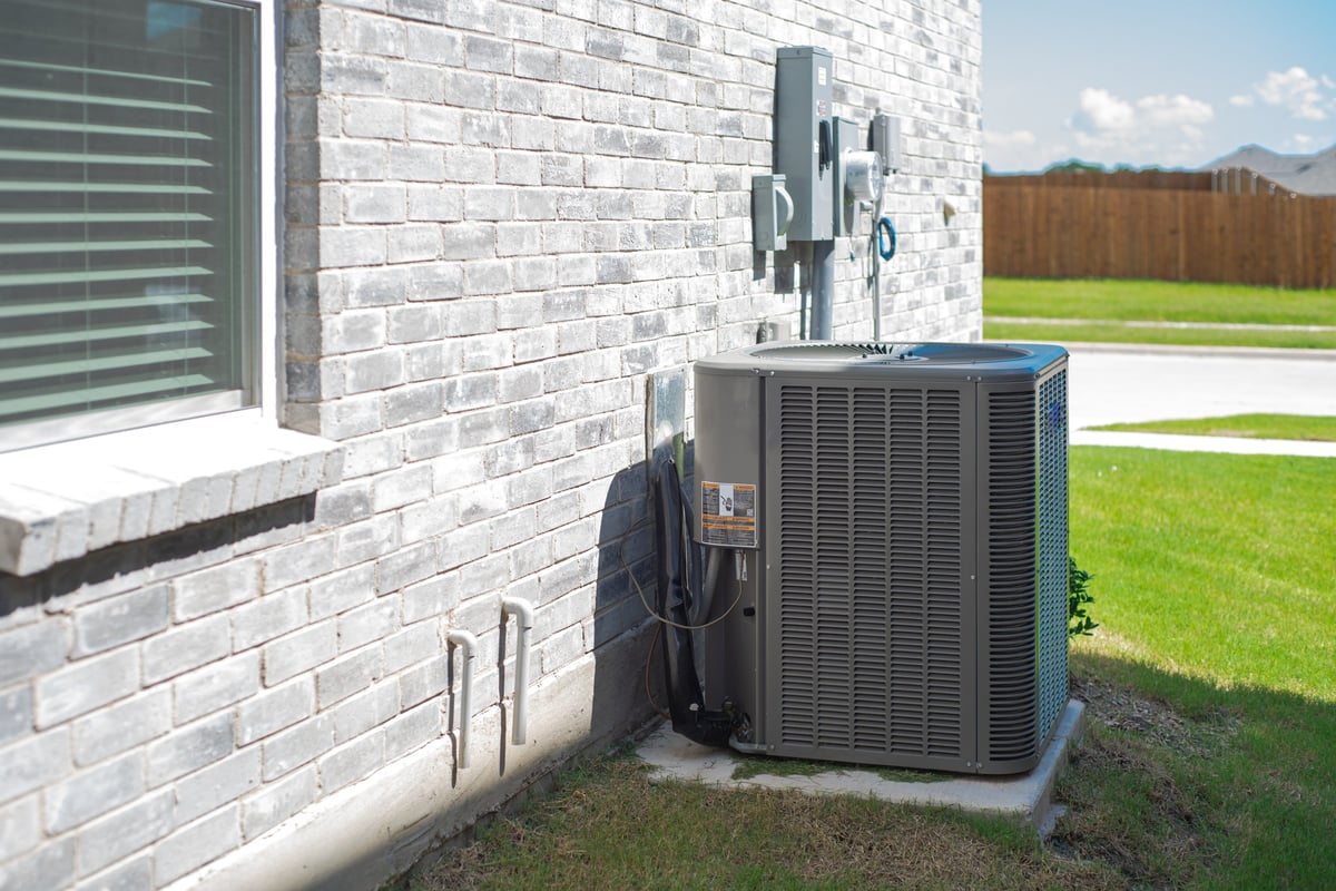 Side windows of new construction home and central air conditioning condenser unit mounted on concrete slab beside brick house wall