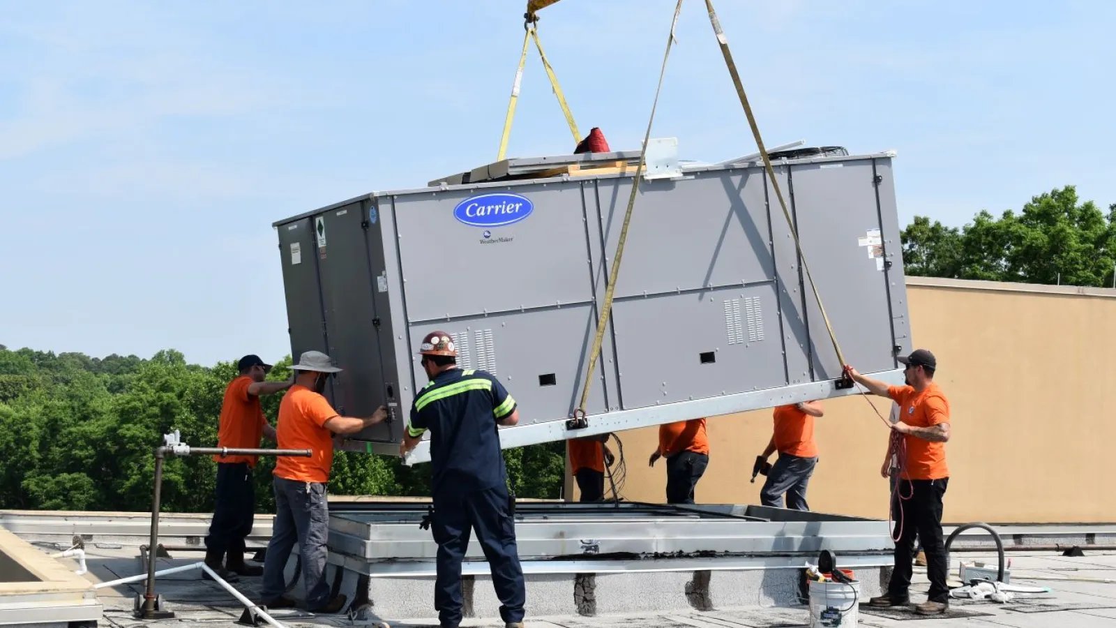 Workers in safety gear using a crane to lift a large Carrier HVAC unit onto a commercial rooftop